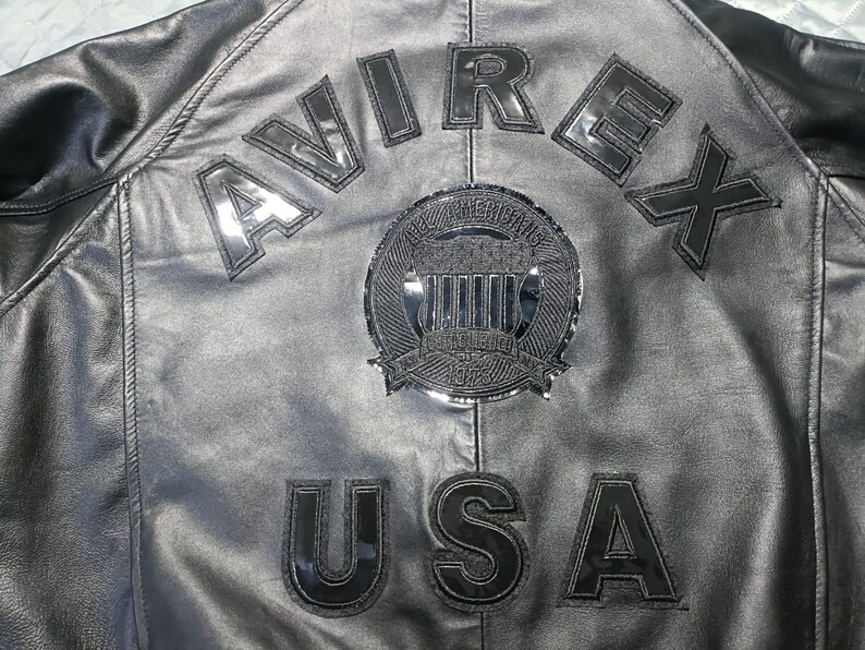 May include: Close-up of a black leather jacket with the word "AVIREX" arched across the upper back. Below the word "AVIREX" is a circular emblem and the word "USA". The jacket has a shiny, reflective finish.
