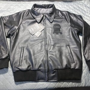 May include: A black leather jacket with a zip-up front, two front pockets, and a ribbed collar and cuffs. The jacket has a patch on the left chest and a tag attached. The jacket is displayed on a light blue quilted surface.