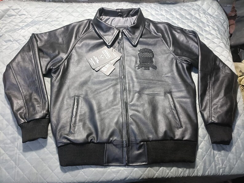 May include: A black leather jacket with a zip-up front, two front pockets, and a ribbed collar and cuffs. The jacket has a patch on the left chest and a tag attached. The jacket is displayed on a light blue quilted surface.