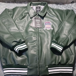May include: A dark green leather jacket with a collared neckline and a zipper closure. The jacket features white and green striped cuffs and hem. An embroidered patch with a patriotic design is on the chest. The jacket has a tag attached.