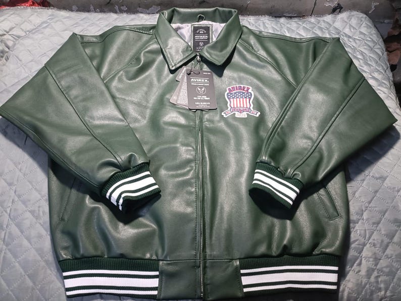 May include: A dark green leather jacket with a collared neckline and a zipper closure. The jacket features white and green striped cuffs and hem. An embroidered patch with a patriotic design is on the chest. The jacket has a tag attached.