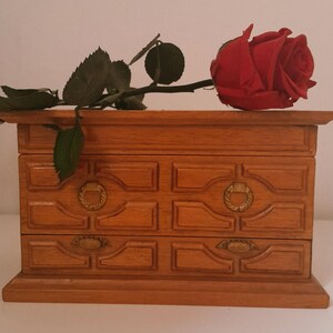 Può includere: Un portagioie in legno con due cassetti e ferramenta decorativa color oro. Una rosa rossa con foglie verdi è appoggiata sopra la scatola, creando un'estetica classica e romantica.