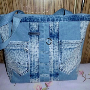 Puede incluir: Un bolso tote confeccionado en tela de mezclilla azul claro con paneles de estampado de leopardo en gris claro. El bolso incluye dos bolsillos exteriores, una franja vertical central con una anilla metálica y una etiqueta que dice handmade.