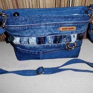 May include: A denim crossbody bag with a long adjustable strap. The bag is made from various shades of blue denim and features a decorative ruffle detail. A small leather tag reads "hand made".