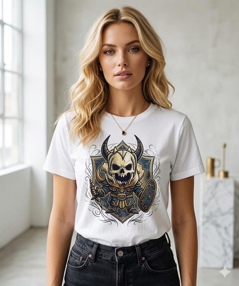May include: White t-shirt with a cartoon illustration of a horned skull warrior, holding a sword and shield. The warrior is in blue, gold, and white, set against a blue and gold shield design.