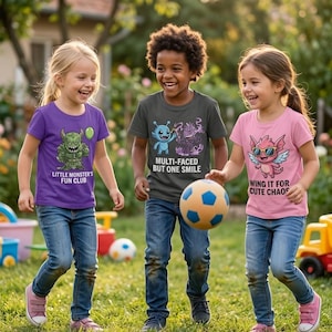 May include: Three children in a garden, wearing graphic t-shirts and jeans, play with a football. The shirts are purple, grey, and pink, each with a monster design and text: "Little Monster's Fun Club", "Multi-Faced But One Smile", and "Wing It For Cute Chaos".