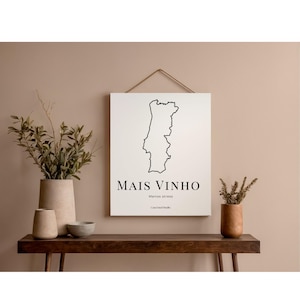 Op de afbeelding: Een witte canvas print met een zwarte omtrek van Portugal, de tekst "MAIS VINHO" en "Menos stress". De print hangt boven een houten tafel met vazen en planten. De achtergrond is een neutraal beige.
