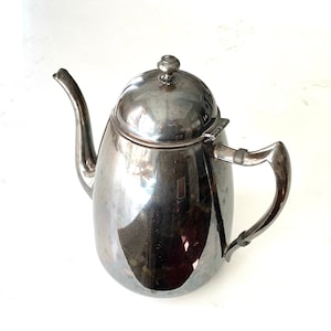 May include: A silver-colored teapot with a curved spout and a rounded body. The teapot has a lid with a small knob and a handle on the side. The teapot is made of a shiny metal and is sitting on a white surface.