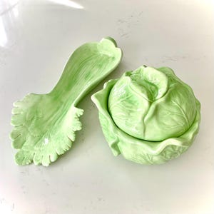 May include: A light green ceramic cabbage-shaped serving dish with a lid, and a matching leaf-shaped serving spoon. The set is designed to resemble fresh produce. The dish and spoon are placed on a white surface.