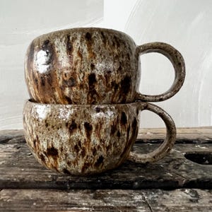 Pode incluir: Duas canecas de cerâmica empilhadas com um esmalte salpicado em tons de castanho, bege e creme. Cada caneca tem um corpo arredondado e uma pega curva no lado direito. A superfície da cerâmica tem um acabamento brilhante com gotas escuras.