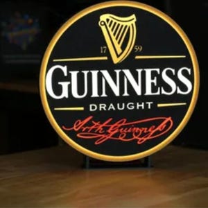 Puede incluir: Un letrero circular iluminado de Guinness Draught con fondo negro, un icono de arpa dorada, el texto "GUINNESS" en blanco, "DRAUGHT" en letras blancas más pequeñas y la firma roja "Arthur Guinness" en la parte inferior.