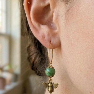 Puede incluir: Un pendiente de aro dorado con una cuenta redonda de color verde moteado, rematada con pequeños detalles metálicos dorados, que cuelga sobre un dije de abeja de latón con acabado antiguo. El pendiente cuelga de un fino aro de hilo dorado.