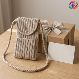 Può includere: Una borsa a tracolla in uncinetto beige con trama a coste, chiusura a patta con bottone dorato e tracolla intrecciata. La borsa è posizionata accanto a una scatola regalo quadrata in cartone con nastro abbinato e un cartoncino bianco.