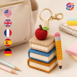 May include: A crochet keychain featuring a stack of three small books in tan, blue, and brown, topped with a red apple with a green leaf. A yellow pencil with a pink eraser is attached to the side. The keychain has a gold metal ring and clasp.