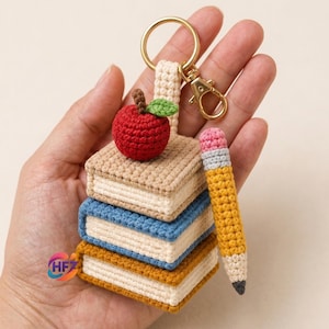 May include: A crochet keyring featuring a stack of three small books in beige, blue, and yellow, topped with a red apple with a green leaf. A yellow pencil with a pink eraser and grey ferrule is attached to the side, connected by a gold ring.