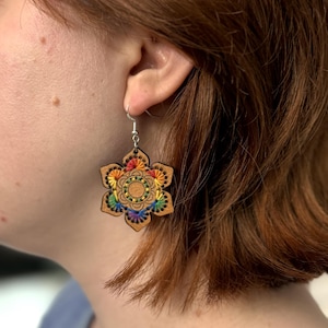 May include: A hexagonal wooden earring featuring intricate laser-cut details and vibrant rainbow-colored embroidery thread stitched in a circular pattern. The earring hangs from a silver-toned metal hook and displays a natural light brown wood grain.