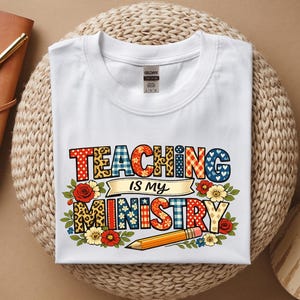 Może przedstawiać: Biała koszulka z napisem "TEACHING IS MY MINISTRY" w kolorowych, wzorzystych literach. Projekt zawiera kwiatowe akcenty, ołówek i transparent. Koszulka jest złożona na tkanej powierzchni.