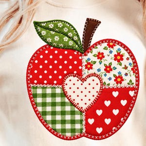 Op de afbeelding: Een crèmekleurig t-shirt met een grote, centrale appelvormige applicatie. De appel is opgebouwd uit vier verschillende stoffen: rood met witte stippen, groen-wit gingham, rood met witte hartjes en een bloemenprint.