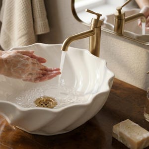 May include: A white, scalloped-edge ceramic sink with a gold faucet. Water streams from the faucet onto hands covered in soap. A clear glass soap dispenser with a gold pump and a bar of soap sit on the wooden counter.