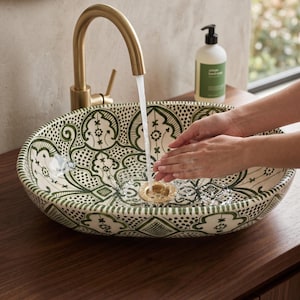 May include: An oval ceramic sink with a green and white patterned design. A gold tap is running water into the sink. A bottle of soap sits on the counter. The sink is on a wooden surface.