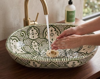 May include: An oval ceramic sink with a green and white patterned design. A gold tap is running water into the sink. A bottle of soap sits on the counter. The sink is on a wooden surface.