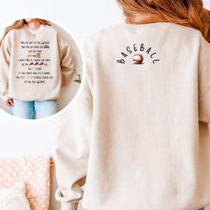 May include: Beige sweatshirt with baseball graphics. The front has text: "Take me out to the game..." and the back has the word "Baseball" arched above a baseball graphic. A casual, comfortable top for sports enthusiasts.