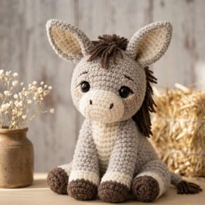 May include: A handmade crochet donkey plush toy in shades of gray, cream, and brown. The donkey has large, expressive eyes, long ears, and a small brown mane. The plush is sitting on a wooden surface, with a vase of flowers and a bale of hay in the background.