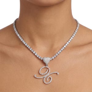 May include: A silver-toned necklace with a heart-shaped pendant and a cursive letter "G" pendant, both encrusted with sparkling clear crystals. The necklace is a tennis style, with a row of clear crystals. The necklace is worn around the neck.