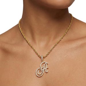 May include: A gold-colored rope chain necklace with a pendant in the shape of the letter "A". The pendant is encrusted with small, clear gemstones. The necklace is displayed on a person.