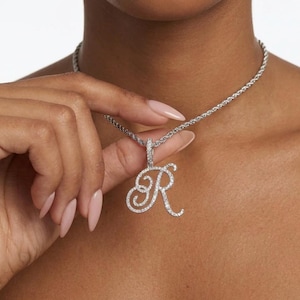 May include: Silver-toned necklace with a pendant in the shape of the letter "R". The pendant is encrusted with small, clear gemstones. The necklace chain is a twisted rope design. The jewelry is displayed against a light background.