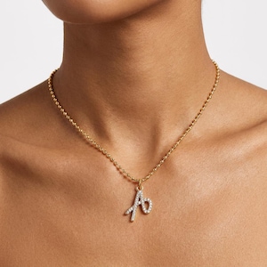 May include: A gold-toned ball chain necklace featuring a script letter A pendant. The pendant is encrusted with small, clear, sparkling stones that cover the entire surface of the letter, creating a bright and reflective appearance against the metal.