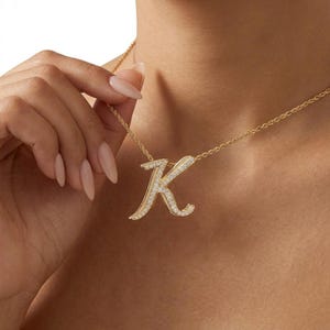 May include: A gold-toned necklace featuring a cursive letter K pendant. The pendant is embellished with small, clear, sparkling stones set along the surface of the letter. The necklace is attached to a thin, twisted gold-toned metal chain.