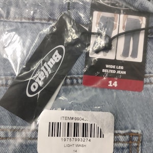 May include: Light wash denim jeans with a wide leg belted design. A black Buffalo Jeans brand tag is attached, featuring a barcode, item number 99043, and a size 14 label. The garment is encased in clear plastic packaging with visible product details.