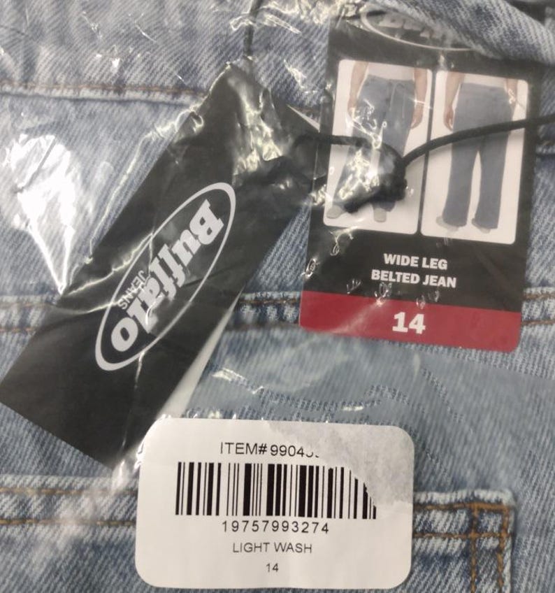 May include: Light wash denim jeans with a wide leg belted design. A black Buffalo Jeans brand tag is attached, featuring a barcode, item number 99043, and a size 14 label. The garment is encased in clear plastic packaging with visible product details.