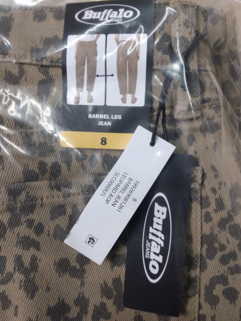 May include: Tan denim jeans with a dark brown leopard print pattern. The jeans feature a Buffalo Jeans brand tag with the text BARREL LEG JEAN and size 8. A secondary white tag displays the text 199269081261 BARREL JEAN LEOPARD AOP SLC0060US.