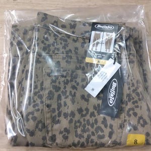 May include: A pair of tan jeans with a dark grey leopard print pattern, enclosed in a clear plastic bag. The jeans feature a Buffalo brand tag, a barrel leg style label, and a size 8 tag. The garment includes a visible back pocket and tags.