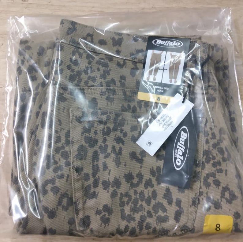 May include: A pair of tan jeans with a dark grey leopard print pattern, enclosed in a clear plastic bag. The jeans feature a Buffalo brand tag, a barrel leg style label, and a size 8 tag. The garment includes a visible back pocket and tags.