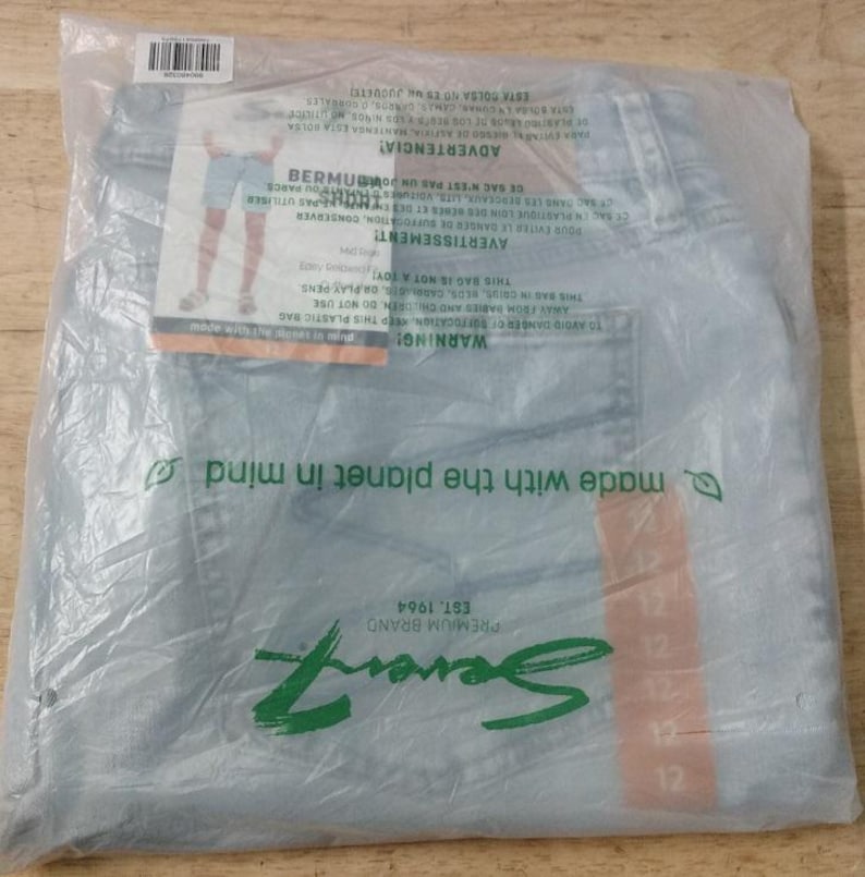May include: Light blue denim shorts packaged in a clear plastic bag. The packaging features the Seven7 Premium Brand logo, a size 12 label, and a graphic showing the shorts being worn. The plastic bag includes safety warnings and environmental text.