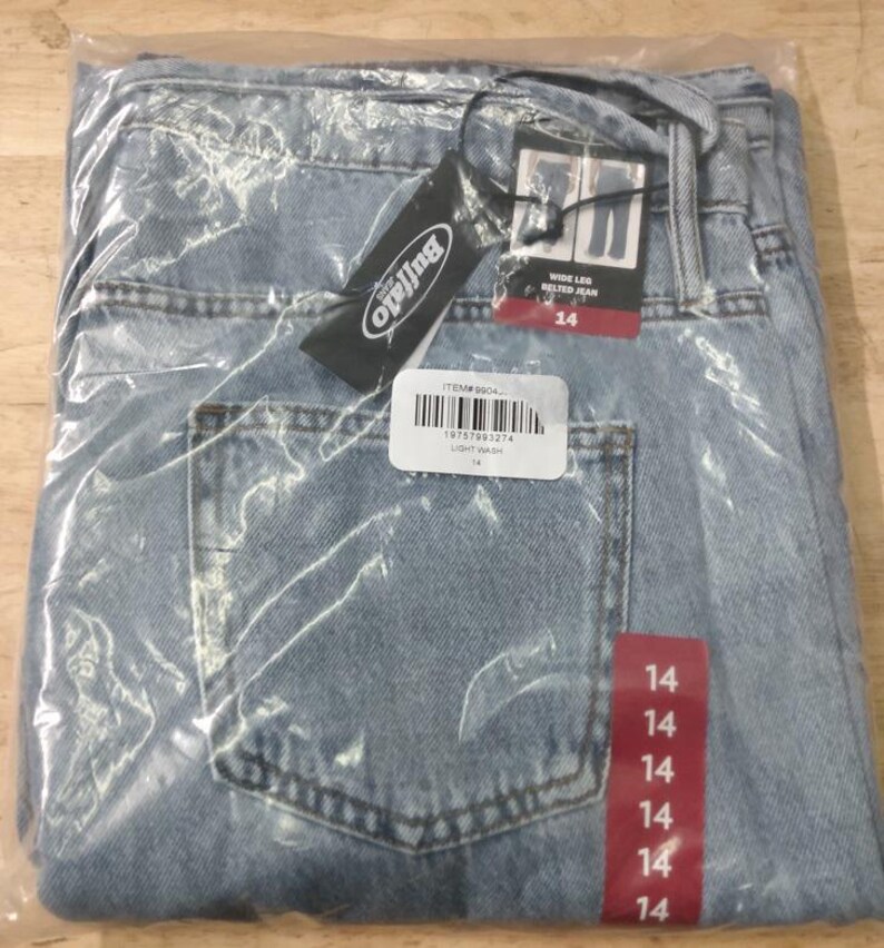 May include: Light wash blue denim jeans in a wide leg style with a belt, enclosed in a clear plastic bag. The jeans feature a Buffalo brand tag, a barcode label with the text ITEM# 9904, and a red vertical strip displaying the size 14 repeatedly.