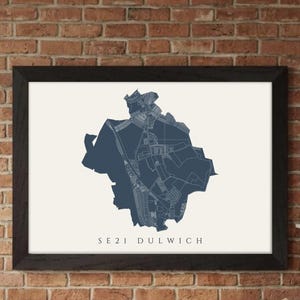 Może przedstawiać: Oprawiony wydruk przedstawiający szczegółową mapę obszaru SE21 Dulwich w ciemnoniebieskim odcieniu na kremowym tle. Mapa jest w czarnej ramie, eksponowana na ceglanej ścianie.