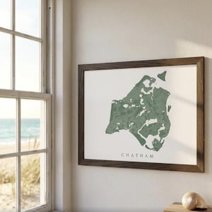 May include: A framed print showing a green map of Chatham on a white background. The map is in a dark brown wooden frame. The word "CHATHAM" is printed below the map. The print is hanging on a white wall, near a window.