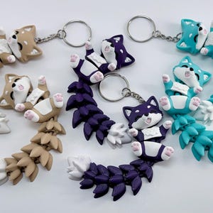 May include: Three articulated fox keychains in tan, dark purple, and teal. Each fox has a white chest, face, and tail tip, with small pink paw pads. The bodies are segmented for movement, and each is attached to a silver-toned metal split ring.