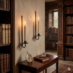 May include: Two black metal wall-mounted candle holders with lit cream-colored taper candles. Below, a dark wood desk holds a circular brass astrolabe, a leather-bound book, a fountain pen, and a small glass inkwell, set against a library background.
