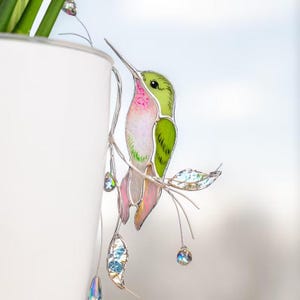 May include: A colorful hummingbird decoration with a long beak, perched on a silver branch with leaves and crystal accents. The bird features green, pink, and white hues. The decoration is near a white pot.