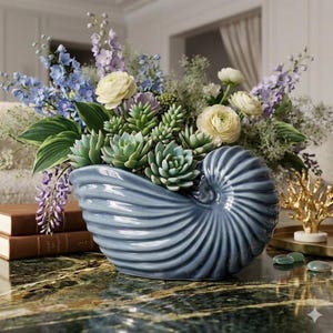 May include: A blue ceramic nautilus shell-shaped vase filled with a variety of flowers and succulents. The arrangement includes blue, purple, and white flowers, and green succulents. The vase sits on a marble surface.