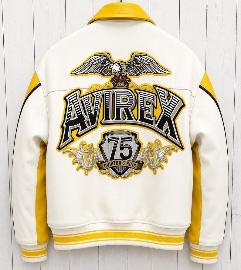 以下が含まれることがあります： 背中に「AVIREX」の文字とワシのグラフィックが入った白と黄色のスタジアムジャンパー。襟、袖、トリムに黄色のアクセント。テキスト「75 Aviator's King」も表示されています。