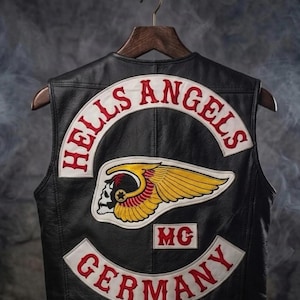 May include: Black leather vest featuring the "Hells Angels" logo in red and white. The logo includes a winged skull and the text "GERMANY". The vest is suspended on a wooden hanger.