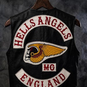 May include: A black leather waistcoat featuring a large embroidered patch on the back. The patch displays the text HELLS ANGELS in red on a white curved background, a central winged skull emblem in yellow and red, the letters MG, and ENGLAND in red.
