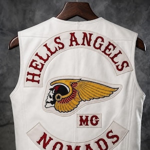 May include: A white leather waistcoat featuring embroidered patches on the back. The top patch reads HELLS ANGELS in red, the centre patch displays a winged skull graphic, a small patch reads MG, and the bottom patch reads NOMADS in red lettering.