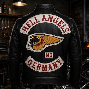 Könnte beinhalten: Eine schwarze Lederjacke mit einem großen Rückenaufnäher, der den Text HELL ANGELS in Rot auf weißem, geschwungenem Hintergrund zeigt, einem zentralen geflügelten Totenkopf-Emblem in Gelb und Rot sowie einem unteren weißen Aufnäher mit rotem Text GERMANY und MC.